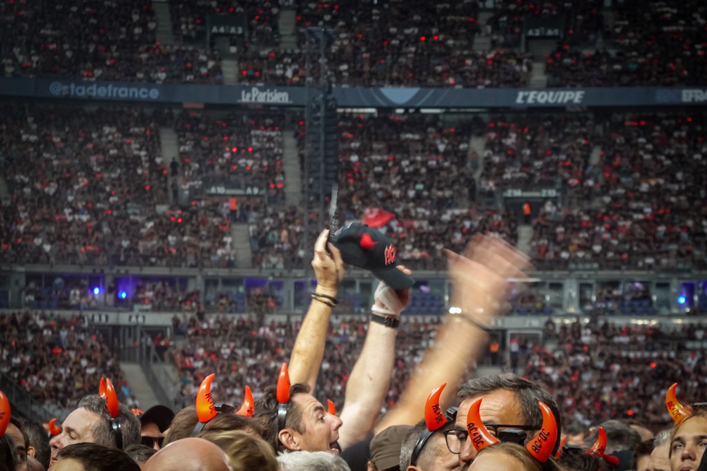 A massive crowd of fans at the Stade de France wearing glowing red AC/DC devil horns during the 2026 Power Up Tour in Paris. The image captures the high-energy atmosphere of the stadium, with one fan holding up a black baseball cap featuring red horns and the AC/DC logo against a backdrop of thousands of spectators.
