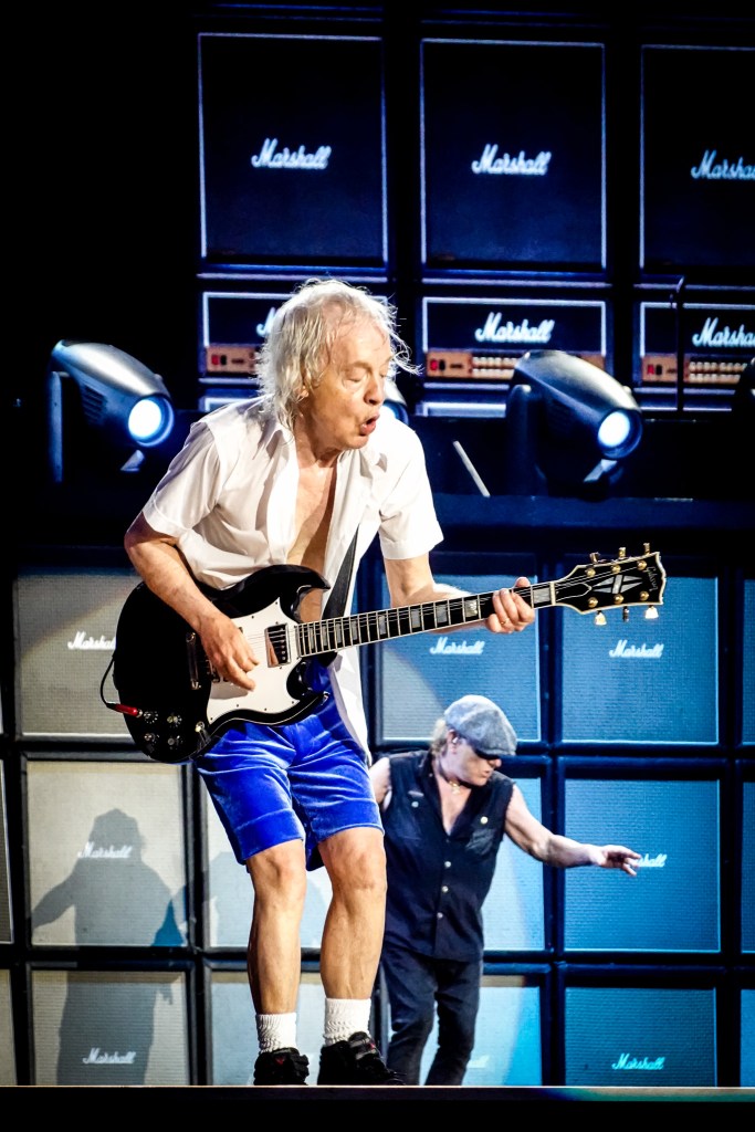 Angus Young leaning into a powerful riff on his Gibson SG during the 2026 Power Up Tour at the Stade de France. He is wearing his blue velvet schoolboy shorts and an open white shirt, performing in front of a towering wall of Marshall amplifiers with Brian Johnson moving in the background under bright stage lights.
