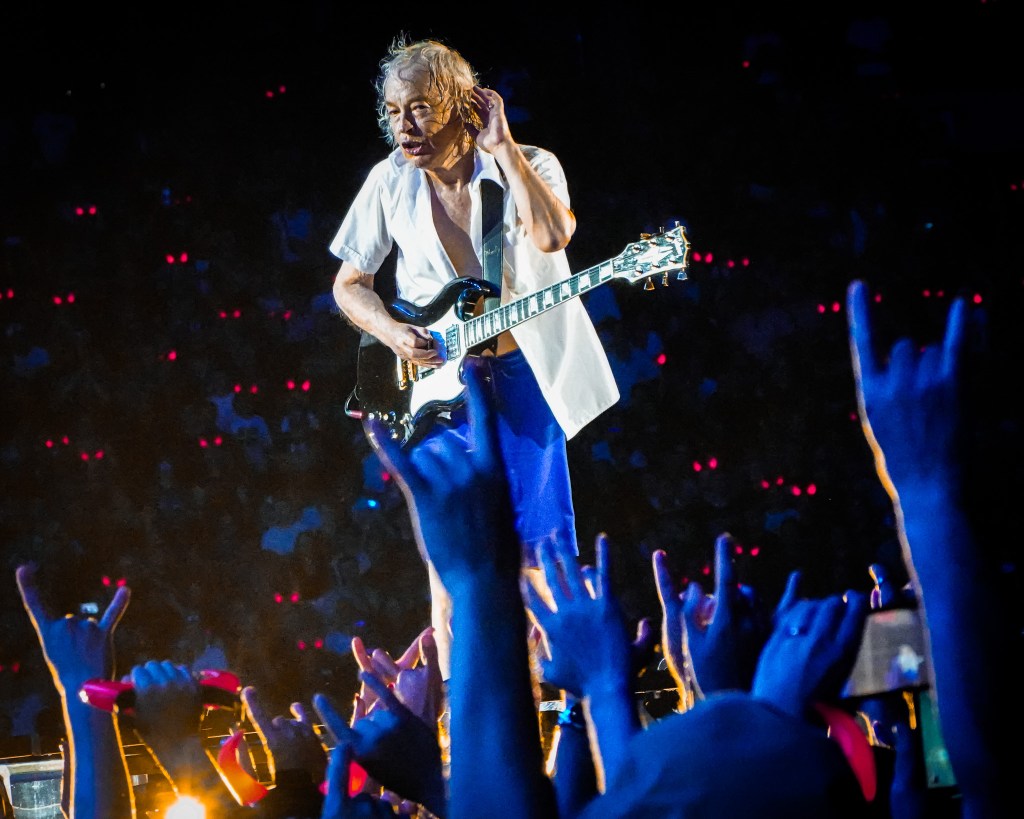AC/DC lead guitarist Angus Young adjusts his hair while holding his black Gibson SG during the 2026 Power Up Tour at the Stade de France. He is wearing his iconic blue velvet schoolboy shorts and an open white shirt, performing above a sea of fans raising their hands in "rock on" gestures, with thousands of glowing red devil horns visible in the dark Parisian crowd.