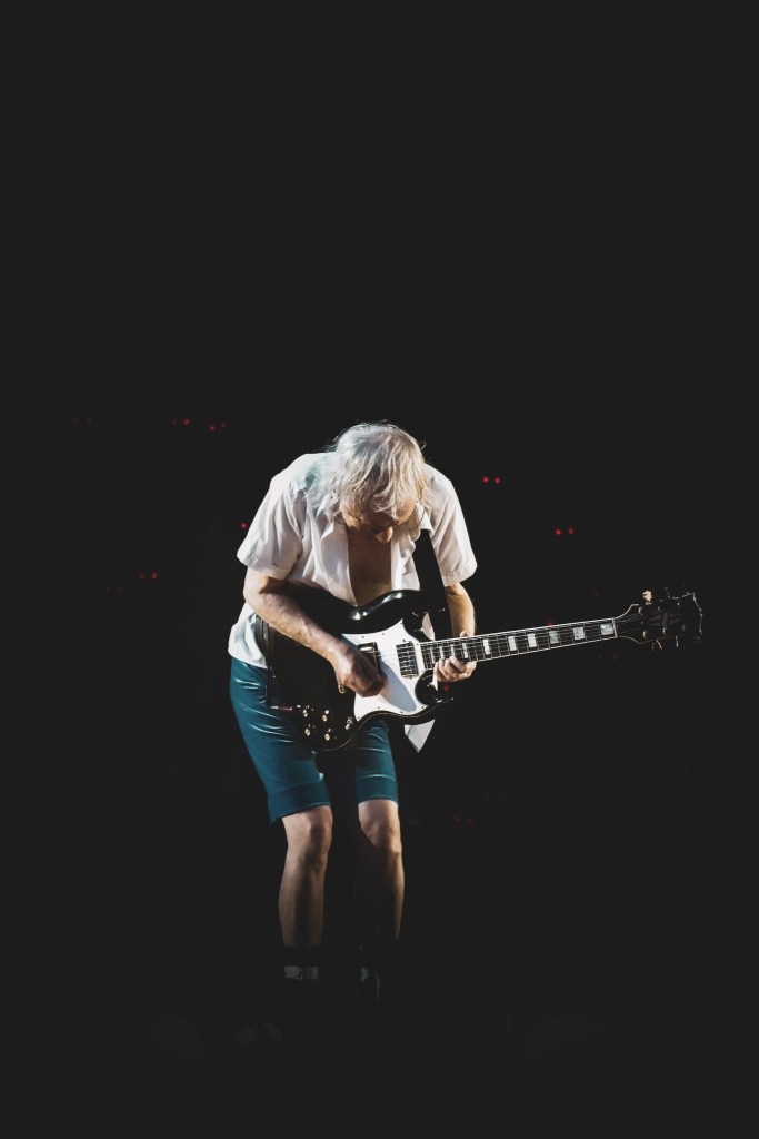 AC/DC lead guitarist Angus Young hunched over his black Gibson SG, lost in an intense solo during the 2026 Power Up Tour at the Stade de France. He is wearing his signature teal velvet schoolboy shorts and an open white shirt, spotlighted against a pitch-black background that emphasizes the raw, focused energy of his performance in Paris.