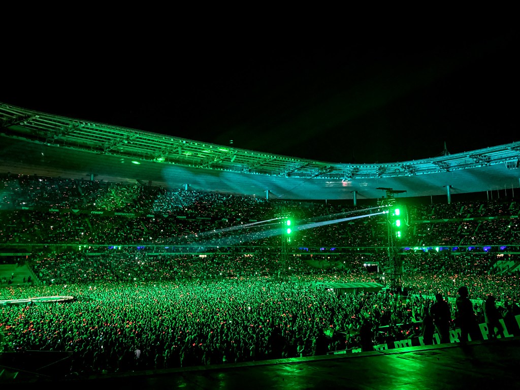 A panoramic view of the sold-out Stade de France during the 2026 Power Up Tour in Paris, with the entire stadium bathed in an electric green glow. The massive crowd is densely packed on the pitch and in the stands, with bright spotlights cutting through the dark night to illuminate the high-voltage atmosphere of the AC/DC concert.