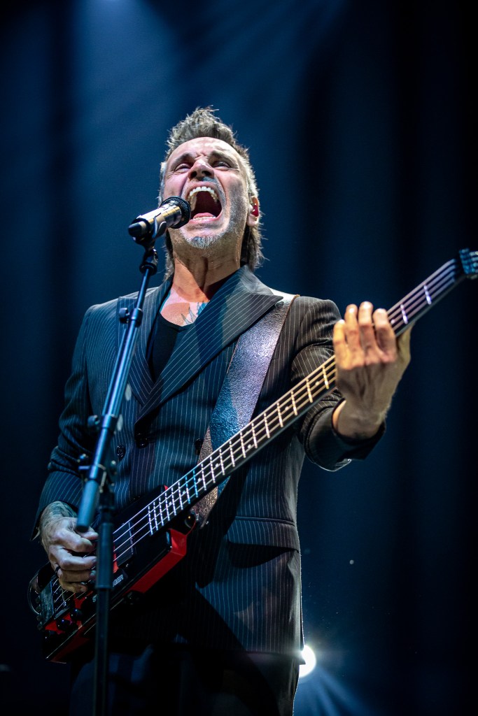 A high-energy, low-angle shot of Italian singer Nek mid-performance, captured while he is singing powerfully into a microphone with his mouth wide open. He is wearing a dark pinstripe suit and playing a red and black headless bass guitar, with a single spotlight from above highlighting his expressive face and spiky hair against a dark background.
