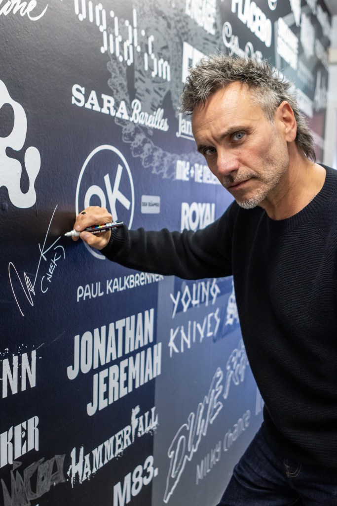 Italian singer Nek looking at the camera while signing a dark wall filled with names and logos of various musical artists, such as Sara Bareilles and Paul Kalkbrenner. He is wearing a dark, textured sweater and using a white marker to add his signature to the collection.
