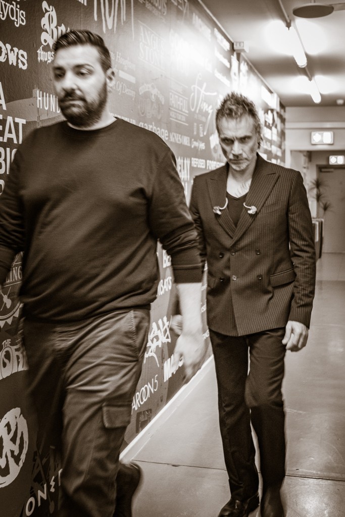 A black and white photograph showing Italian singer Nek walking down a narrow hallway, backstage at a venue. He is wearing a sharp, double-breasted pinstripe suit with in-ear monitors draped around his neck, looking intensely toward the camera. A man in a dark sweatshirt and cargo pants walks slightly ahead of him in the foreground