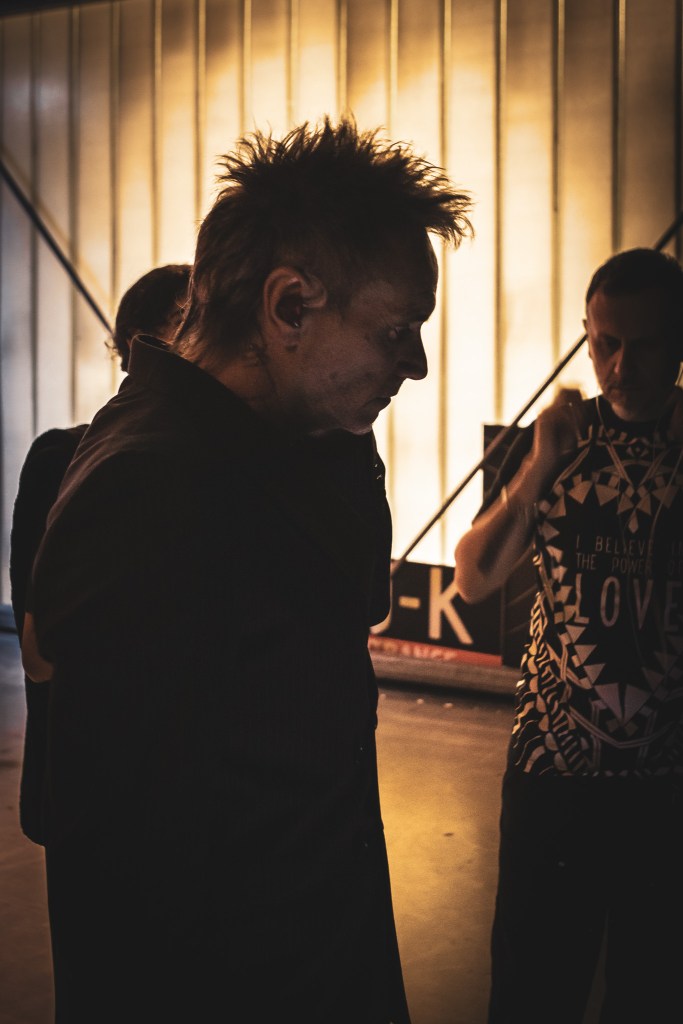 A candid profile shot of Italian singer Nek backstage, looking downward in a moment of quiet concentration. He is wearing in-ear monitors, with his signature spiky hair silhouetted against a warm, vertically slatted light source. Another person is partially visible in the background to the right in the dimly lit environment.