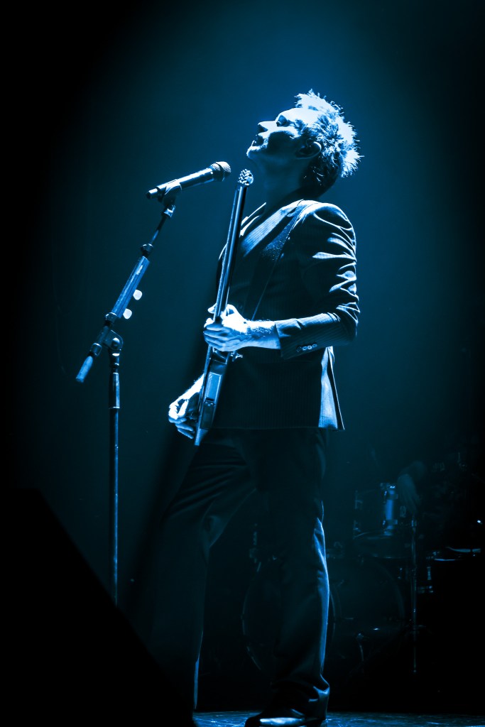 A profile shot of Italian singer Nek on stage, bathed in dramatic blue light. He is looking upwards with a soulful expression, holding a headless bass guitar vertically in front of a microphone stand against a dark background.
