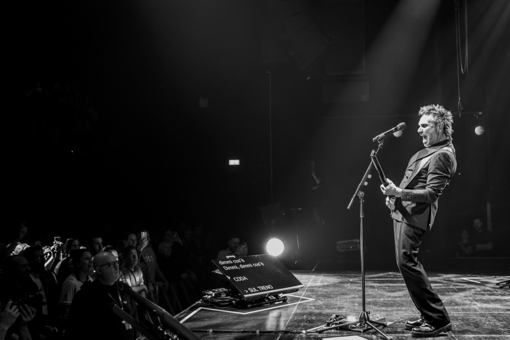 A wide, black and white shot of Nek performing on stage from a profile view. He is singing into a microphone and playing his headless bass guitar, leaning back with high energy. In front of him on the stage floor, a teleprompter displays lyrics in Italian, including the words "Dimmi cos'è" and "SUL TRENO". A portion of the audience is visible in the lower-left corner, watching the performance.
