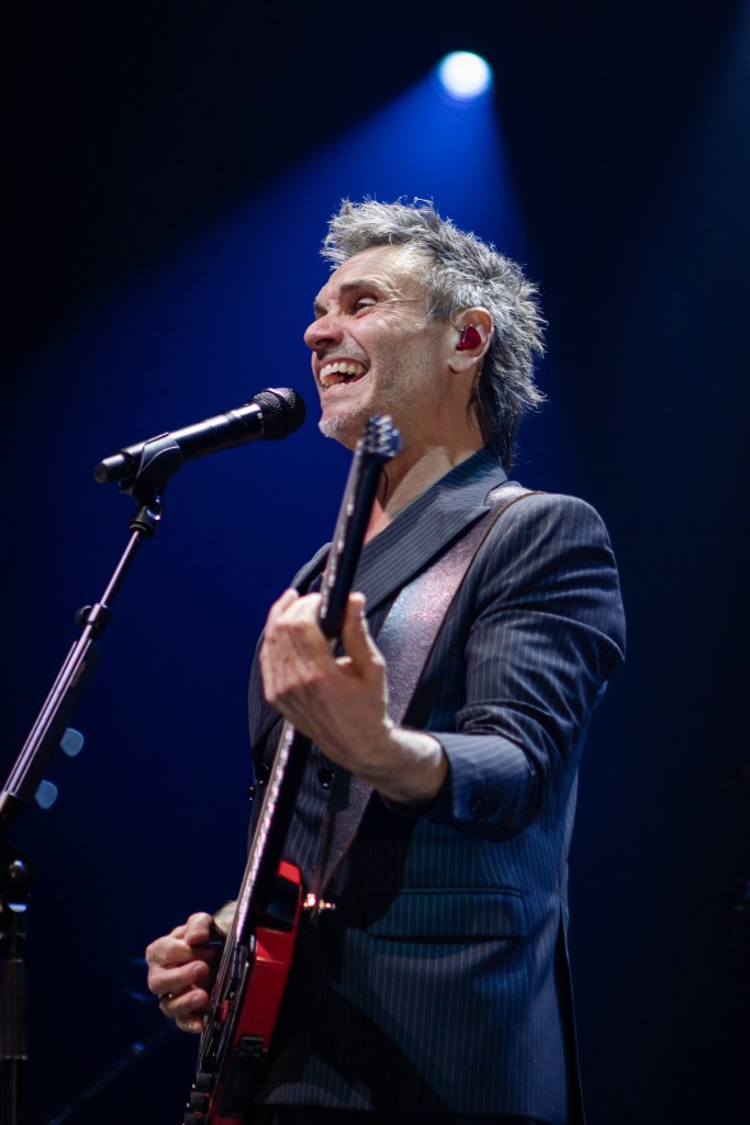 A candid, medium shot of Italian singer Nek smiling broadly while performing on stage. He is captured from a low angle, wearing a dark pinstripe suit and red in-ear monitors. He holds his signature headless bass guitar, positioned in front of a microphone stand, while a single blue spotlight shines from above.