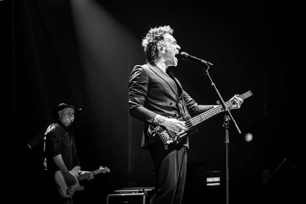 A black and white profile shot of Nek singing passionately into a microphone while playing his headless bass guitar. He is wearing a dark, double-breasted pinstripe suit, with a bright spotlight highlighting him against the dark stage. In the background, another band member in a hat is visible playing a traditional electric guitar.