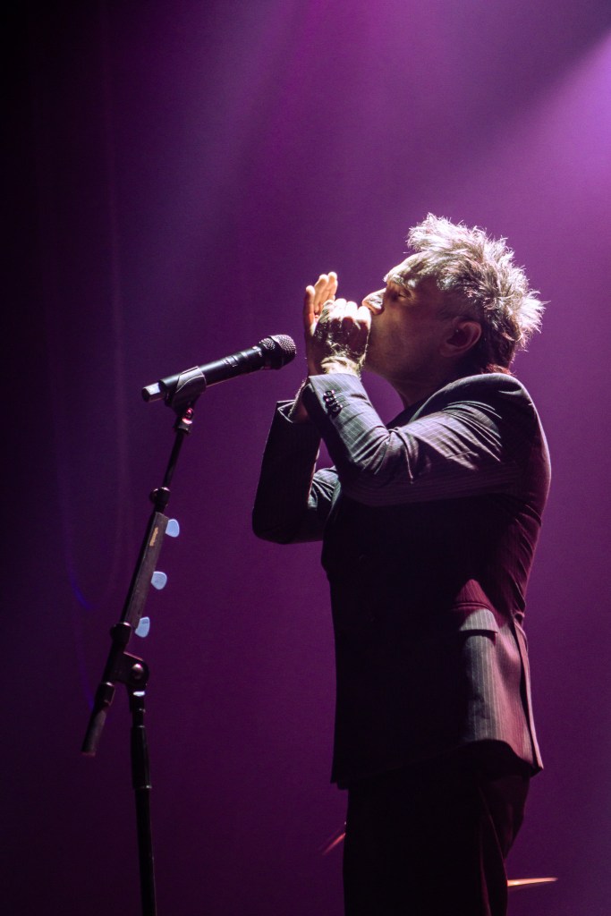 A profile shot of Italian singer Nek performing on stage, bathed in soft purple light. He is seen leaning into a microphone and playing a harmonica, which he holds with both hands. He is wearing a dark, pinstripe suit, and his signature spiky hair is highlighted by the stage lighting.