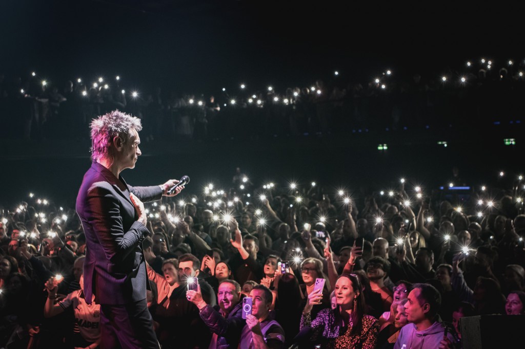 A wide shot from behind and to the side of Italian singer Nek, standing on stage and looking out at a large, cheering crowd. The audience has their phones raised, creating a sea of tiny, bright lights that illuminate the dark venue. Nek is wearing a dark suit and holding a microphone, appearing to take in the moment during a live concert.