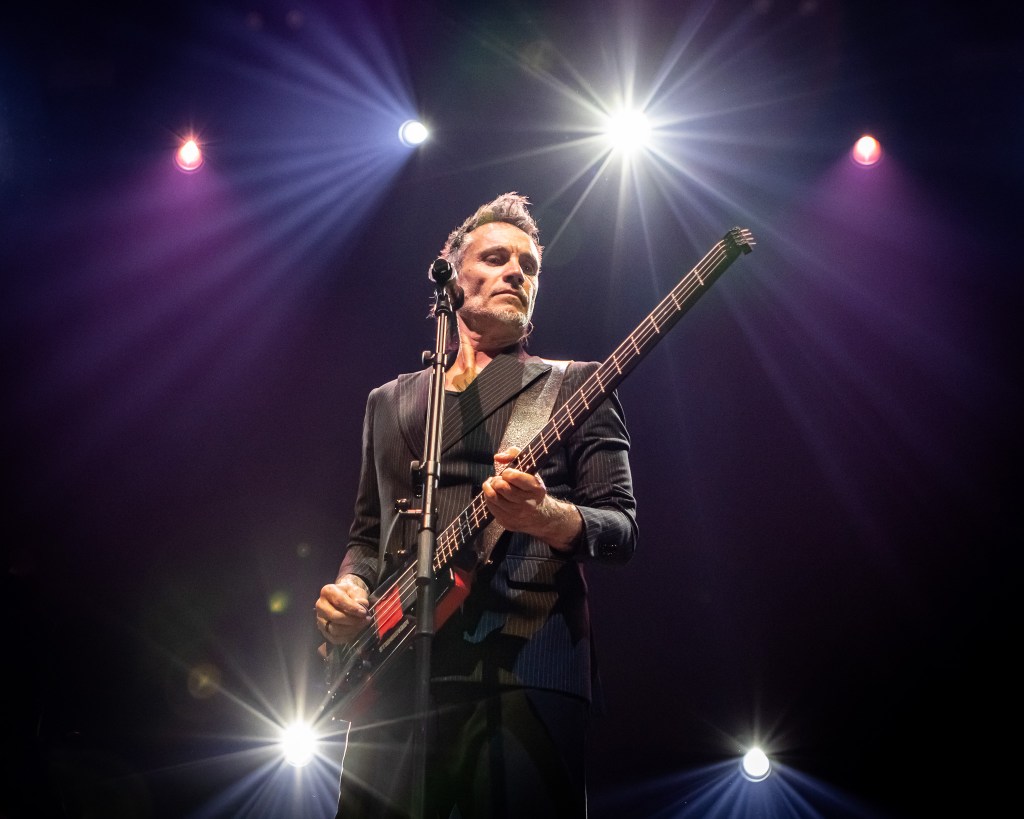 An Italian singer-songwriter, Nek, performing on a stage illuminated by bright spotlights. He is seen from a low angle, wearing a dark pinstripe suit and playing a headless electric bass guitar.