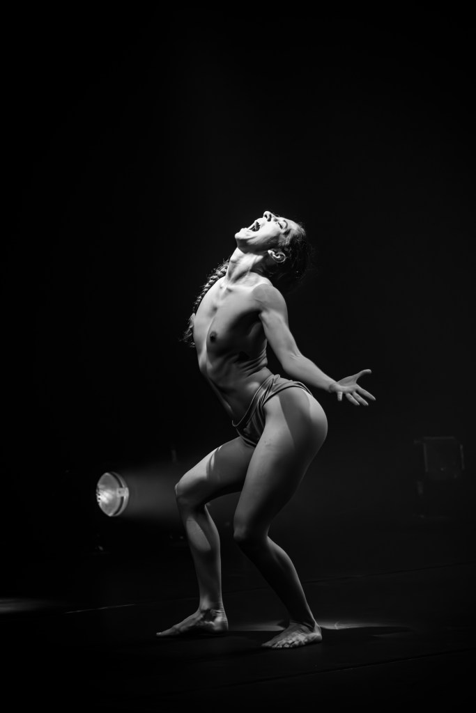 A bare-chested female dancer on a dark stage, illuminated by a low spotlight.: The upper body arches dramatically backward from the waist. Her head is thrown back, facing upward with the mouth wide open in an intense expression or shout.
