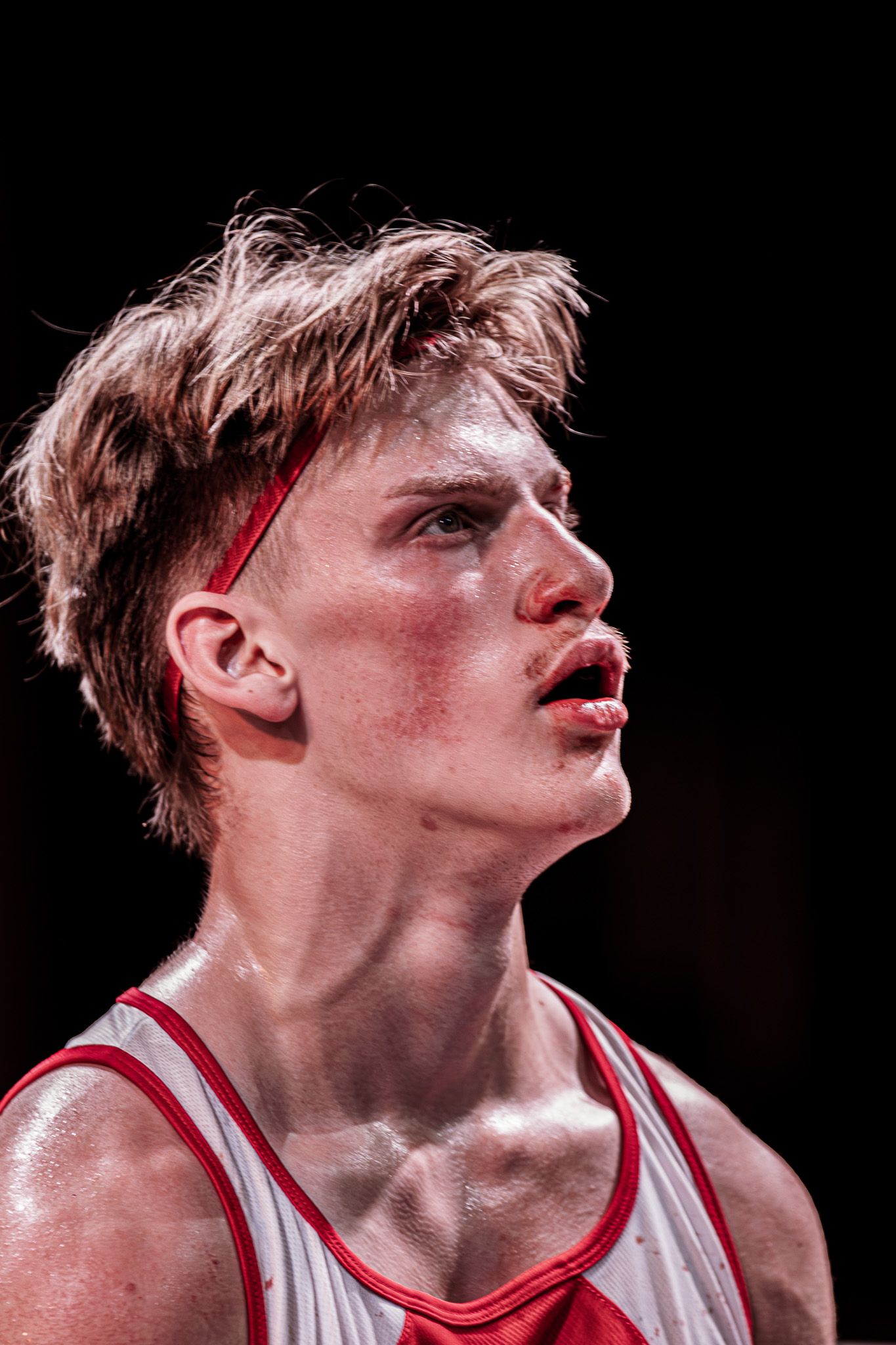 Close-up profile of a focused boxer looking up, wearing a red and white singlet and with blood around his mouth.